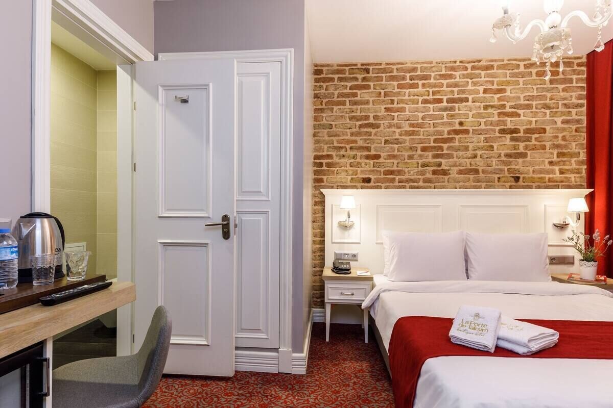 Habitación de hotel con pared de ladrillo, cama con sábanas blancas, escritorio con silla, mesita de noche con lámpara y alfombra roja.