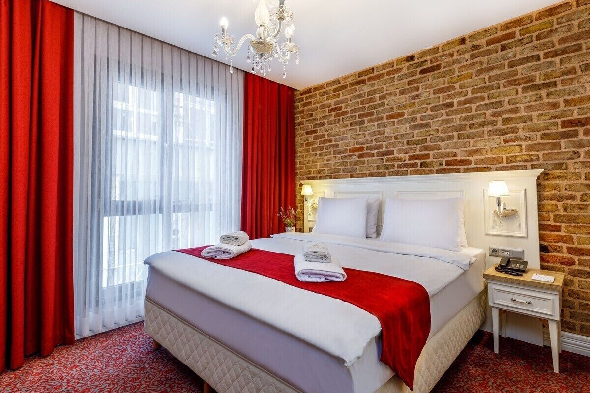 Una habitación de hotel con pared de ladrillo, una cama grande con ropa blanca, cortinas rojas y un candelabro.
