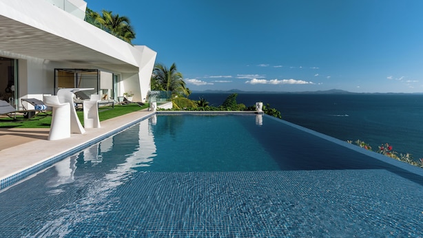 Una piscina moderna con agua azul cristalina, rodeada por un edificio blanco con grandes ventanales y vistas al océano.