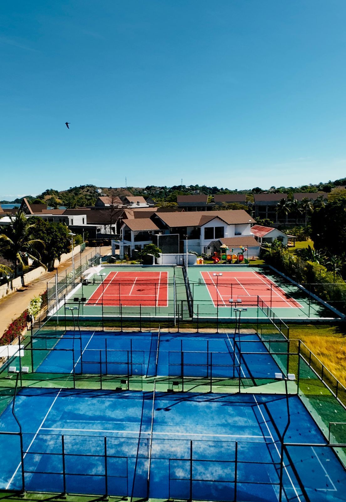 A tennis court complex with multiple courts, surrounded by residential buildings and greenery.