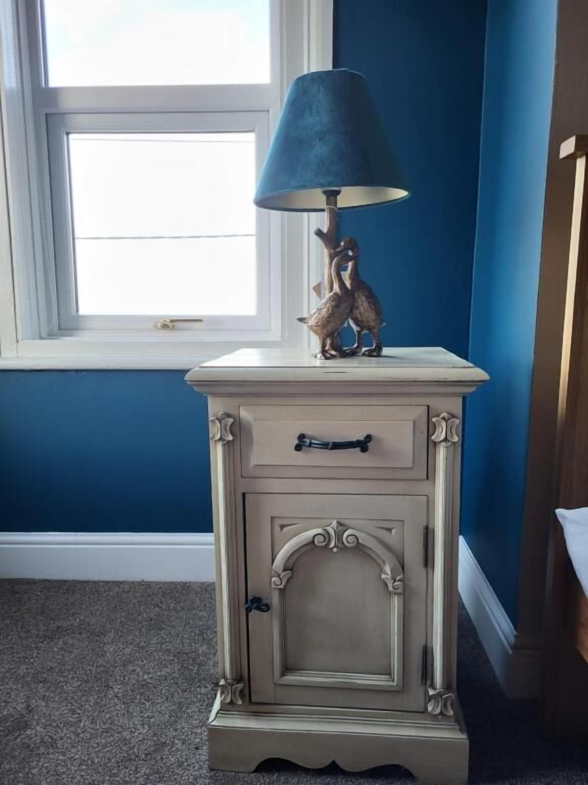 Una mesita de noche de estilo vintage con una lámpara, un cajón y una figura decorativa.