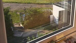 Vista desde una ventana a un paisaje costero con un campo verde, un edificio pequeño y el mar al fondo.