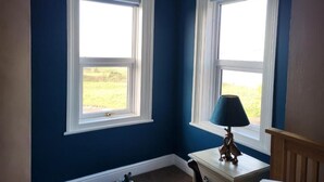 Una habitación con dos ventanas, una mesita con una lámpara y una silla.