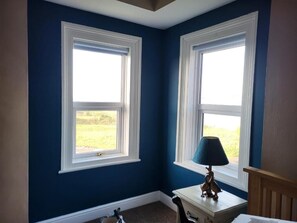 Una habitación con dos ventanas, una mesita con una lámpara y una silla.