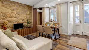 A living room with a stone wall, a sofa, a TV, a dining area with chairs and a table, and a door with an exit sign. - Casa ao Lado – In the Heart of Ponte de Lima with Private Entrance and Full Comfort (Ponte de Lima)