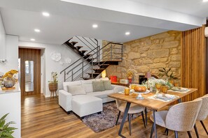 A modern living room with a wooden floor, a white sofa, a dining table with chairs, a staircase, and a stone wall. - Casa ao Lado – In the Heart of Ponte de Lima with Private Entrance and Full Comfort (Ponte de Lima)