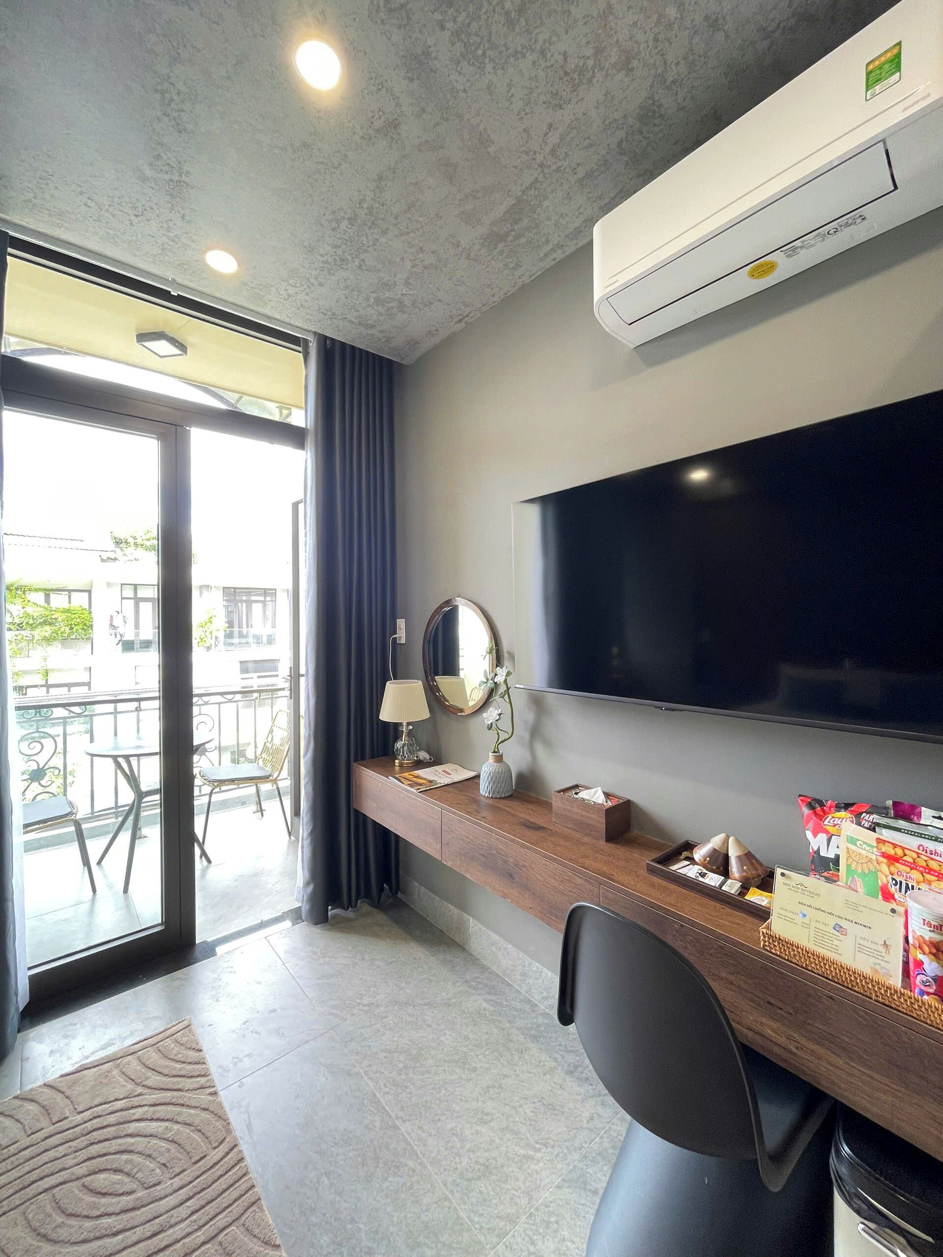 A hotel room with a flat-screen TV, a wooden desk with a lamp, a mirror, and a small plant. There is a balcony with a table and chairs outside.