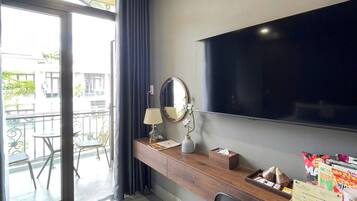 A hotel room with a flat-screen TV, a wooden desk with a lamp, a mirror, and a small plant. There is a balcony with a table and chairs outside.