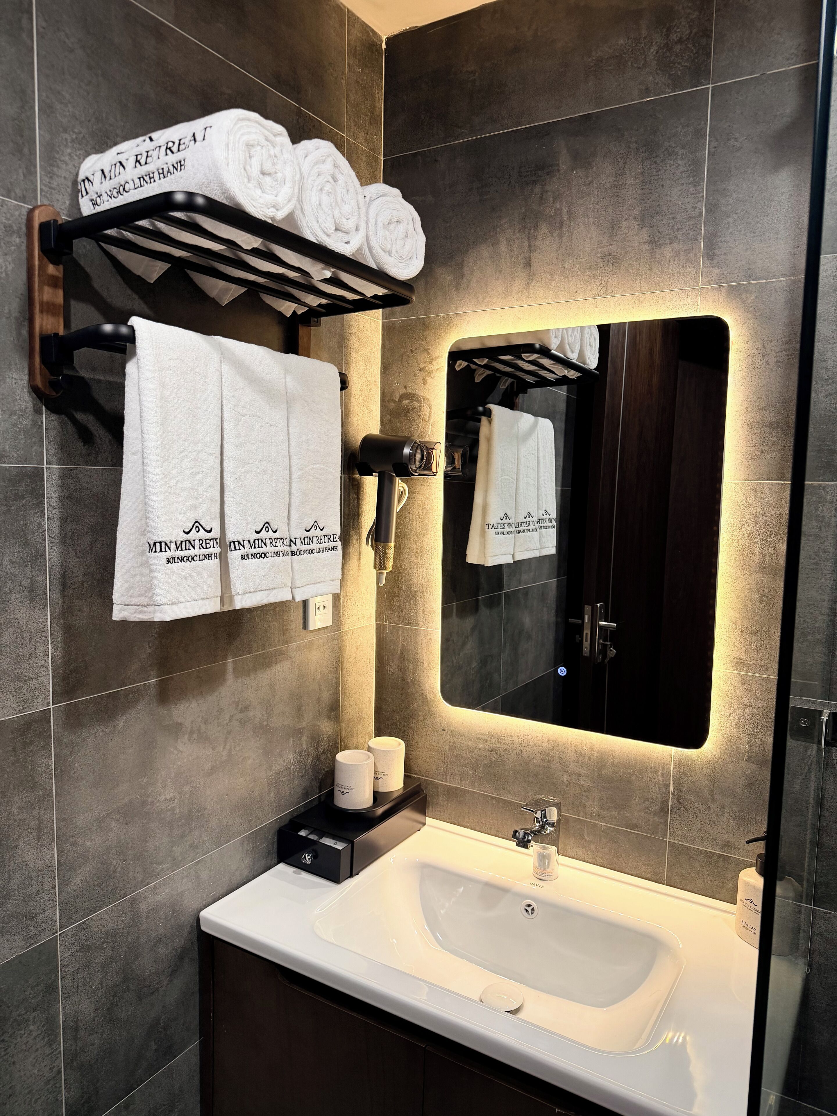Een moderne badkamer met een grote spiegel, een handdoekrek met opgerolde handdoeken, een wastafel en toiletartikelen.