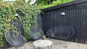 A patio with two black wireframe chairs and a small round table, set against a wooden fence and a hedge. - 4 Star Holiday Home in Skagen (Skagen)