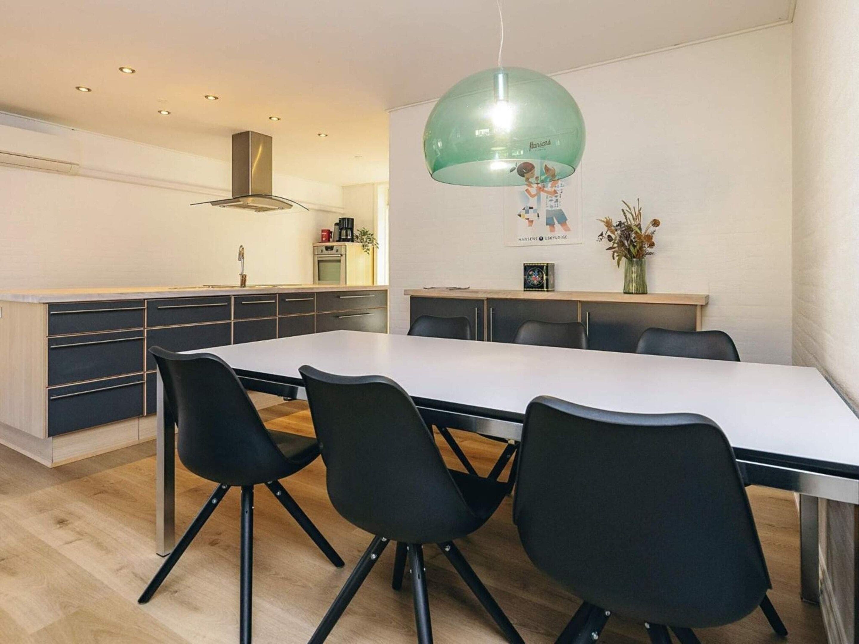 A modern kitchen with a dining area, featuring a long table, black chairs, and a pendant light.