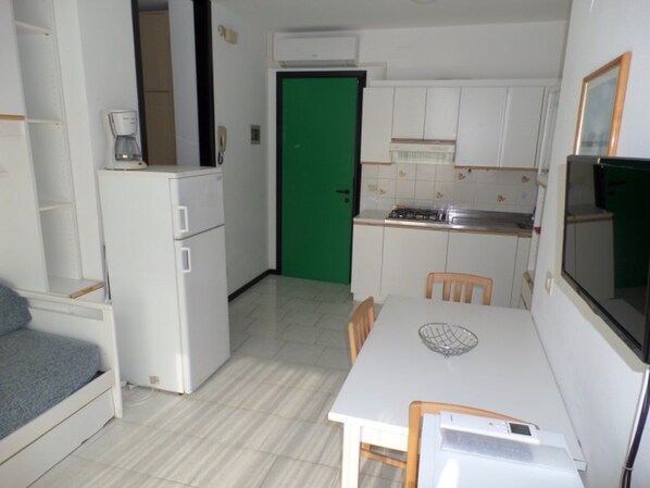 A compact kitchen with white cabinets, a dining table, chairs, a small refrigerator, and a green door. - Apartment near the beach with town views (Bibione)