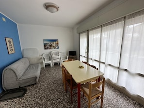 Ein Zimmer mit einer Couch, einem Tisch mit Stühlen, einem Fernseher und einem Fenster mit Vorhängen.