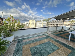 Eine Dachterrasse mit Schachbrettboden, Topfpflanzen, einem weißen Tisch und Blick auf Gebäude im Hintergrund.