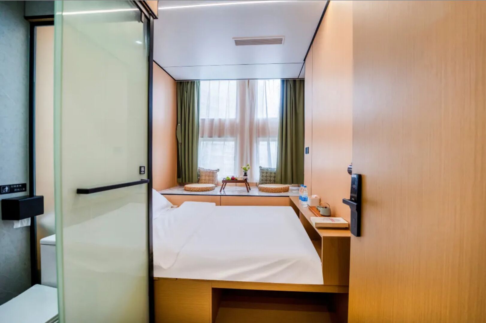 Una habitación de hotel con una cama, una mesita con una botella de agua, una ventana con cortinas y un panel de control en la pared.