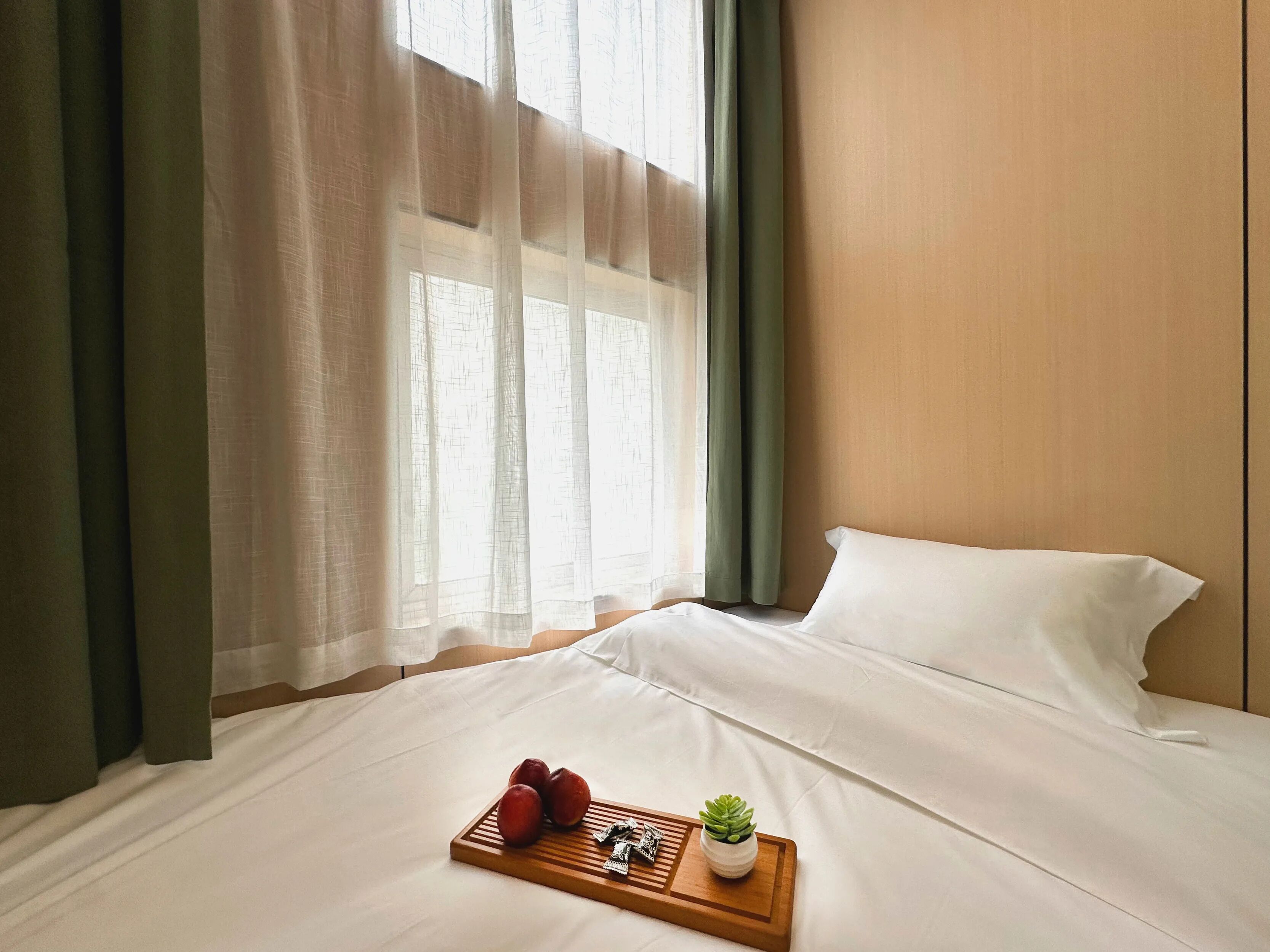 Een hotelkamer met een bed, een houten dienblad met fruit en een plant, en dunne gordijnen.