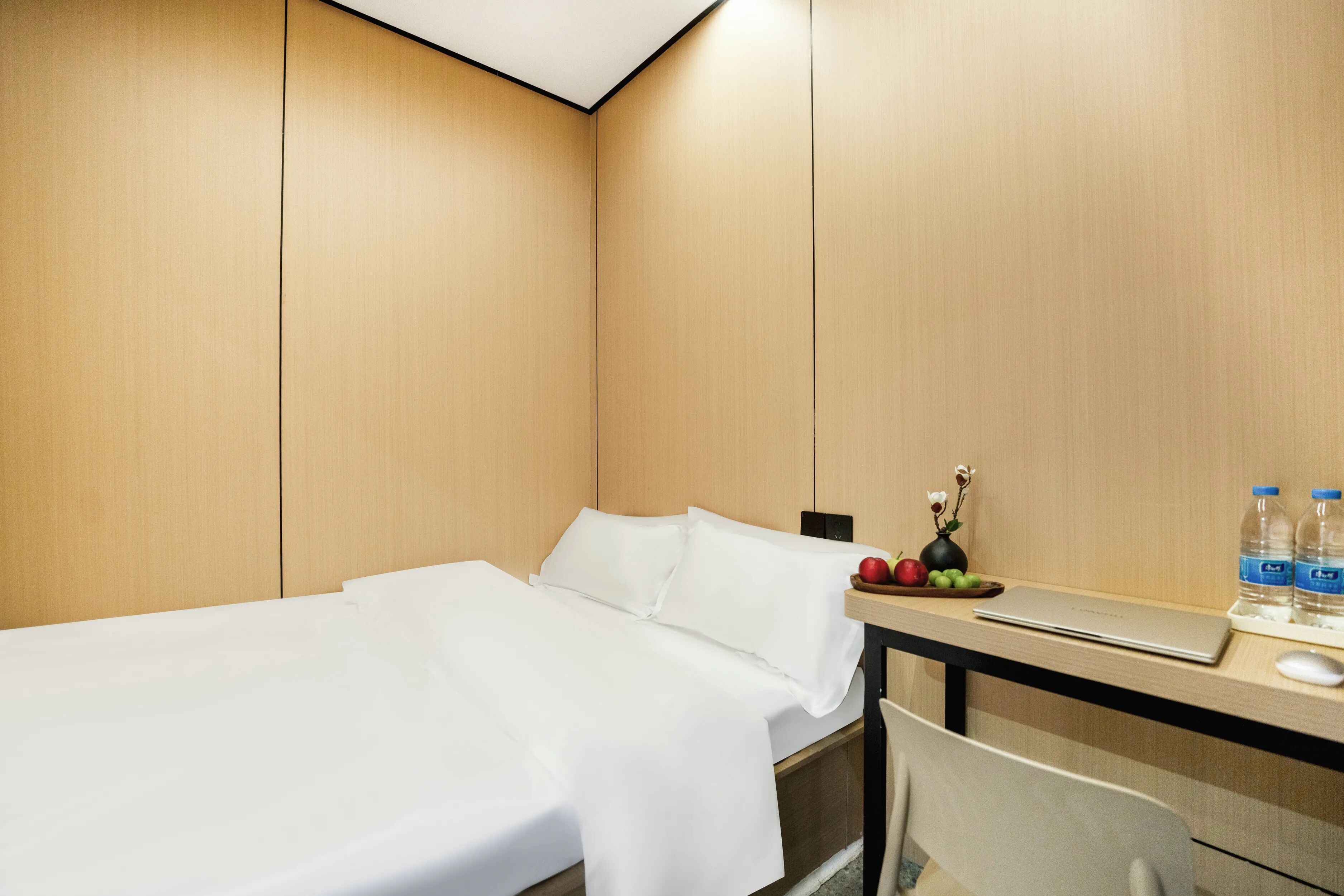 A hotel room with a bed, desk, and a small table with a laptop, water bottles, and fruit.