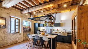 A rustic kitchen with a stone wall, wooden beams, a central island with stools, and a hanging pot rack. - Meublé de tourisme classé 3 étoiles (Fontvieille)