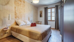 A bedroom with a stone wall, a bed with a beige bedspread, a bedside table with books, and a window with curtains. - Meublé de tourisme classé 3 étoiles (Fontvieille)