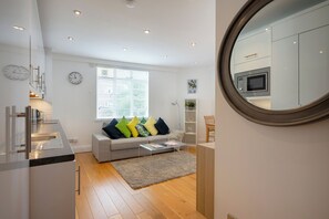 Eine moderne Küche mit integriertem Mikrowellenherd, ein graues Sofa mit farbenfrohen Kissen, ein kleiner Couchtisch und ein großer Spiegel an der Wand.
