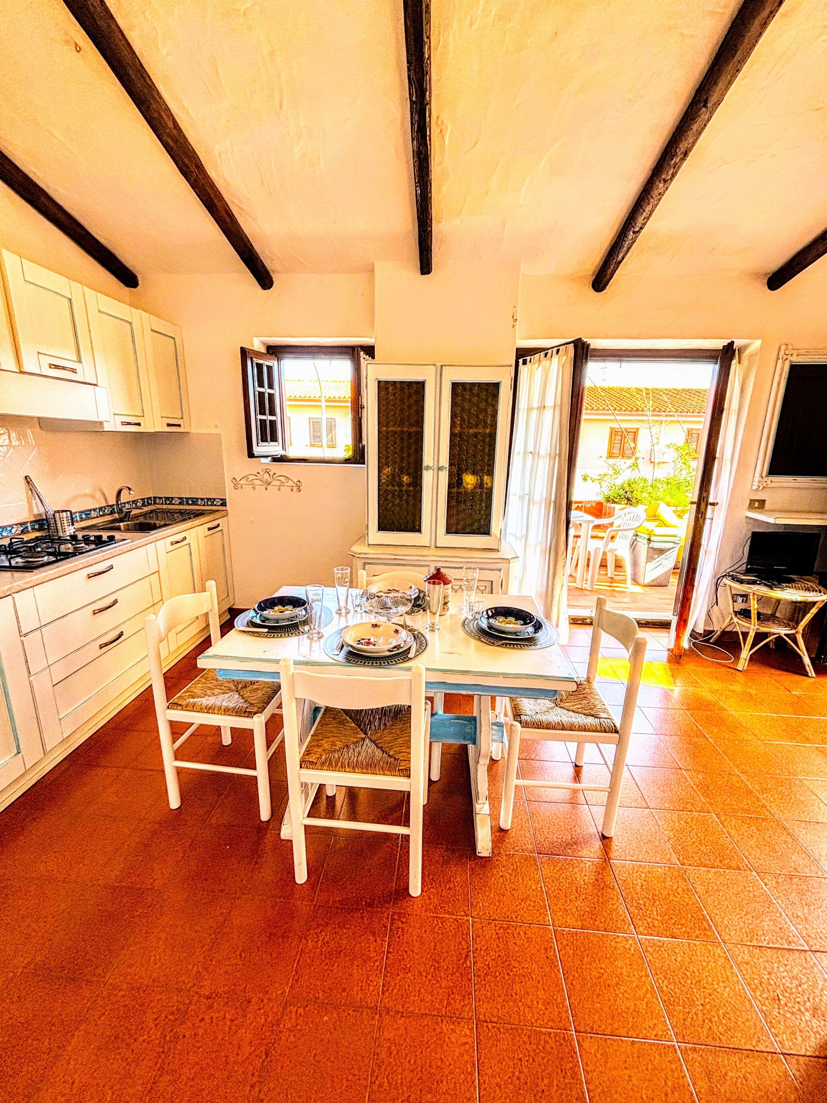 Uma sala de jantar com mesa posta para quatro pessoas, armários de cozinha, fogão e janela com cortinas.