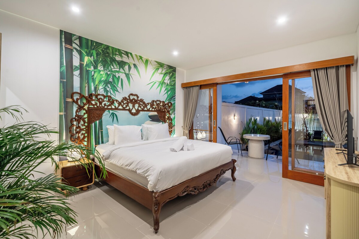 A bedroom with a bed, a wall mural, a sliding glass door to a balcony, and a small table with chairs.