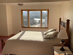 A bedroom with a large bed, a nightstand with a lamp, and a window with a view of a lake and buildings. - 2-Minute Walk to Main Street & Mirror Lake, 2BR Apt overlooking Mirror Lake (Lake Placid)