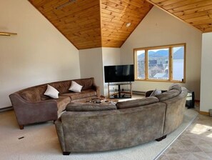 A living room with a large sectional sofa, a flat-screen TV, and a large window with a view. - 2-Minute Walk to Main Street & Mirror Lake, 2BR Apt overlooking Mirror Lake (Lake Placid)