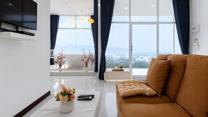 A modern living room with a sofa, a flat-screen TV mounted on the wall, a large window with curtains, and a view of the landscape. - A View Khao Soon  (Chawang)