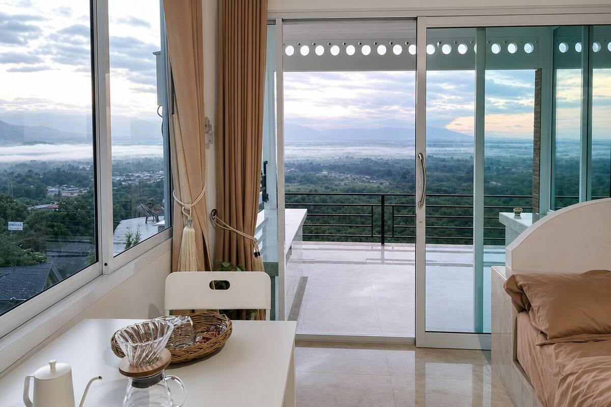 A bedroom with a large window, a bed, a table with a kettle and a glass carafe, and a view of a cityscape and mountains.