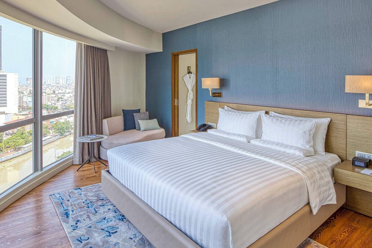 Une chambre d’hôtel dotée d’un grand lit, d’un coin salon avec un canapé et des fauteuils, d’une petite table et offrant une vue sur la ville grâce à de grandes fenêtres.