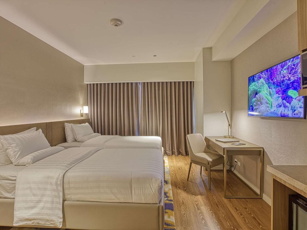 Habitació d'hotel amb llit, escriptori, cadira, televisió, cortines i llum al sostre.