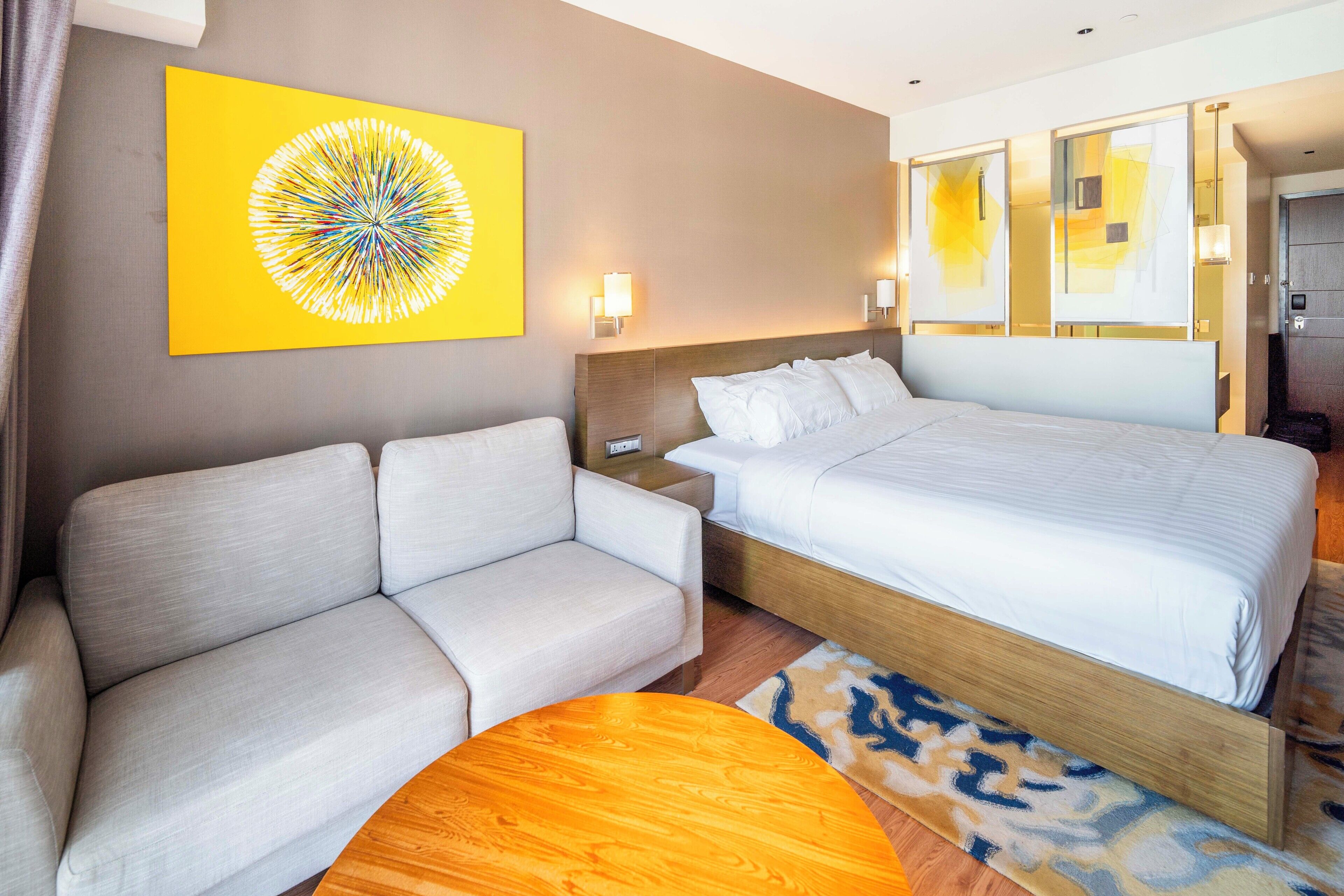 A hotel room with a bed, a sofa, a small wooden table, and a yellow artwork on the wall.