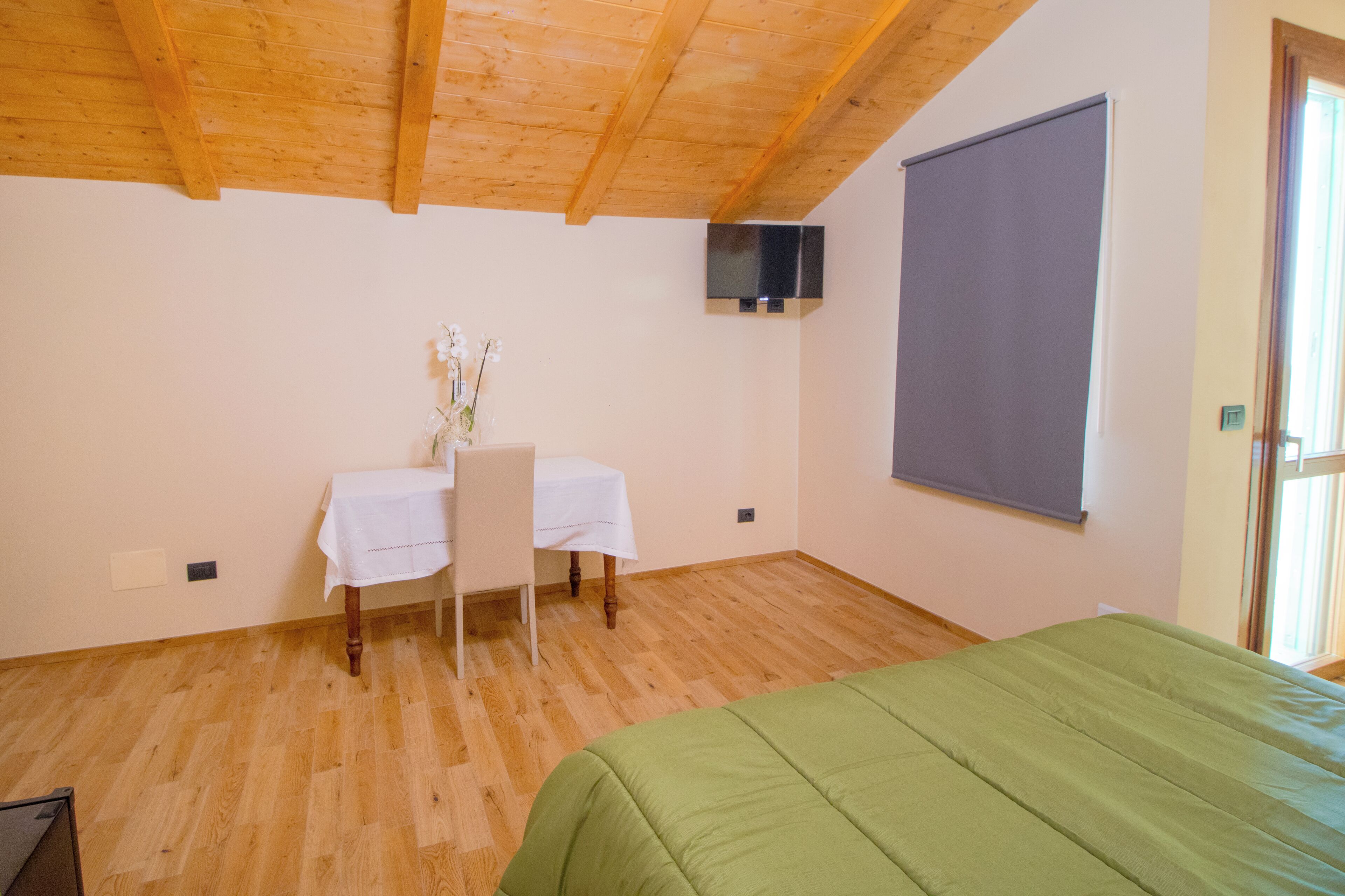 Una habitación con techo de madera, una cama con cubrecama verde, una mesa con mantel blanco, una silla, una planta pequeña y un televisor en la pared.