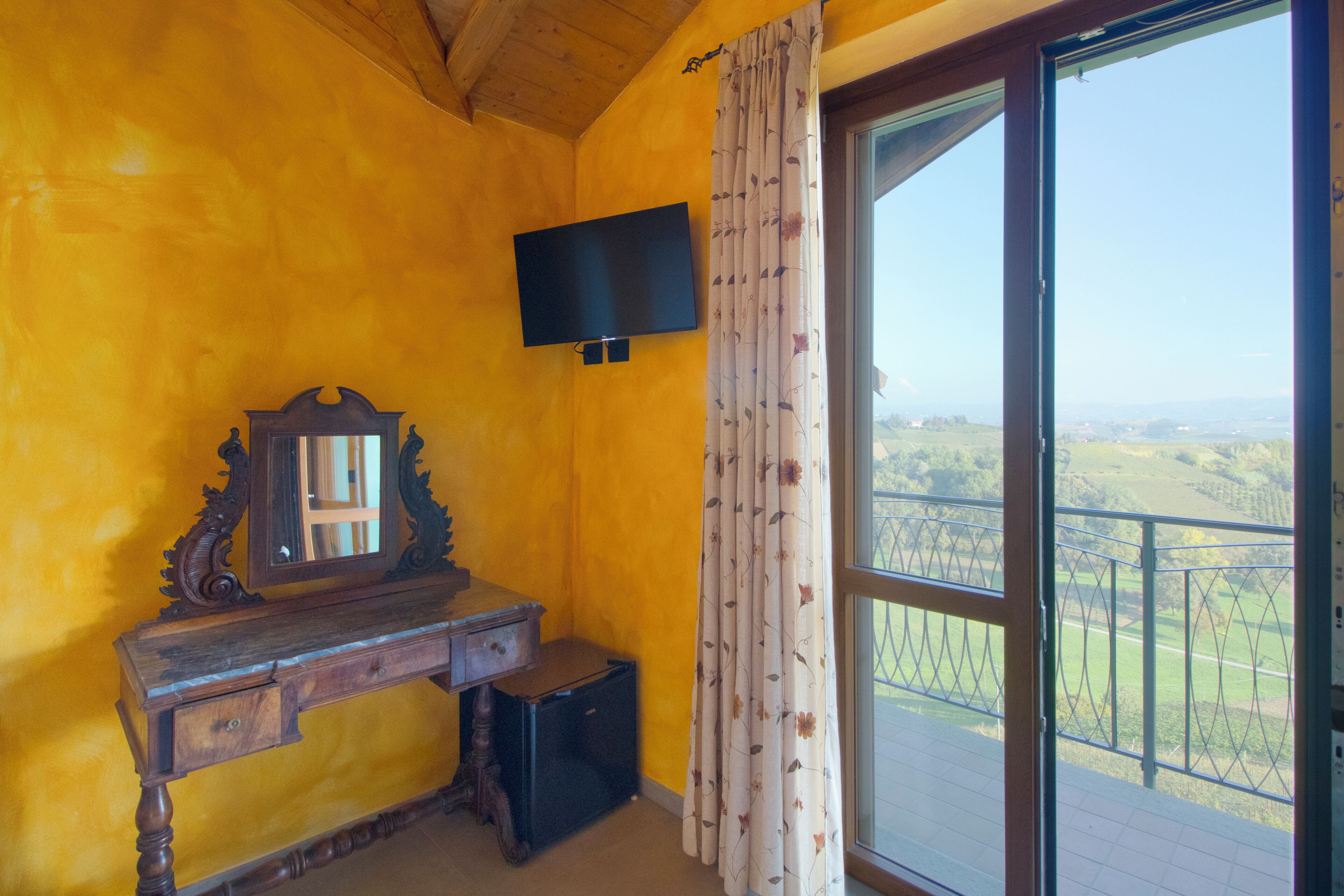 Una habitación con escritorio de madera, espejo, televisión y un amplio ventanal con vistas al campo.