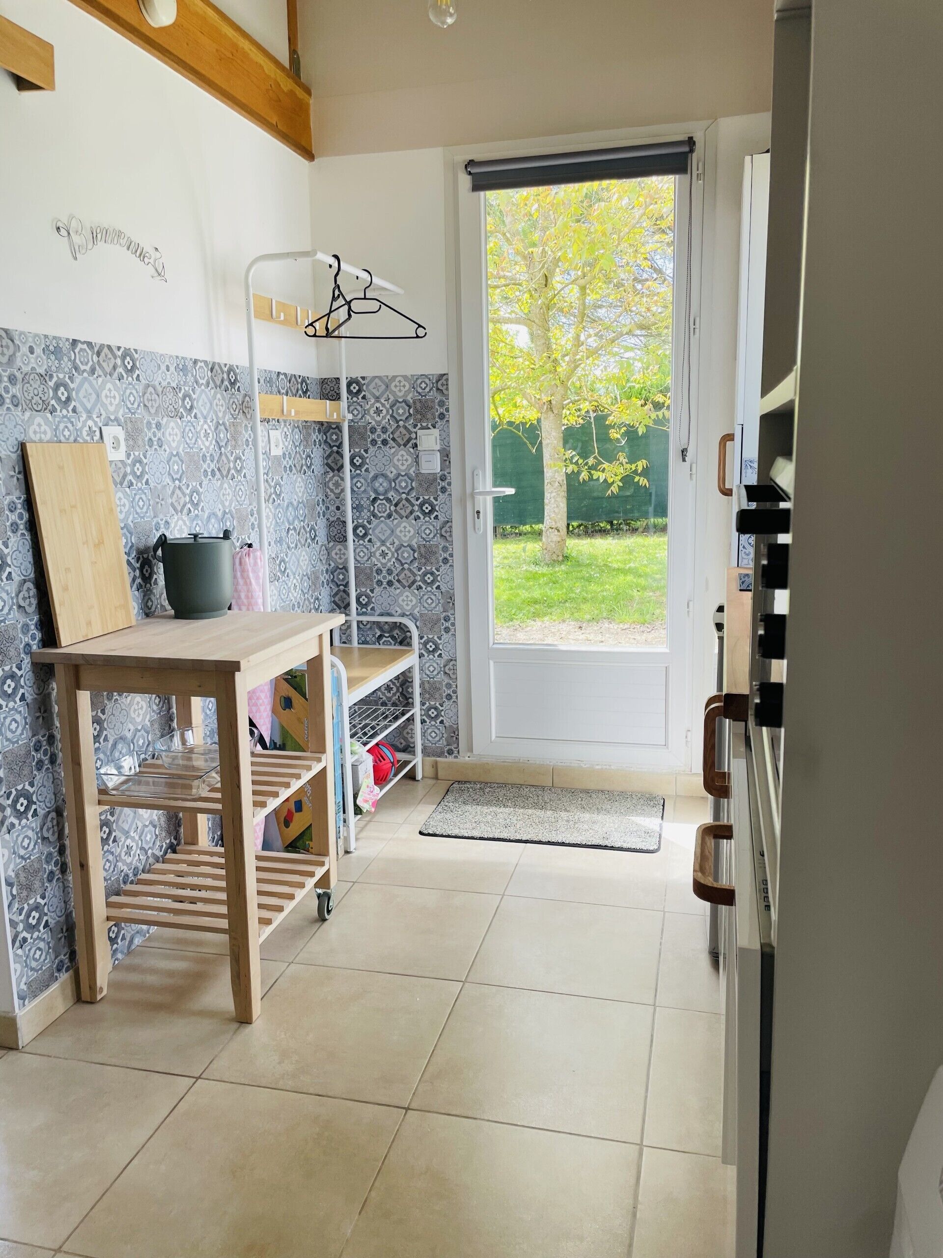 Une cuisine avec une table en bois, un étagère suspendue, un mur recouvert de carreaux et une porte donnant sur l’extérieur.