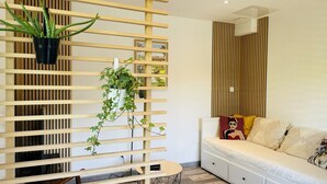 A modern living room with a wooden slatted partition, a sofa with cushions, a small round table, and a hanging plant. - Cosy 3-star house with private garden and quiet terrace (Mézériat)