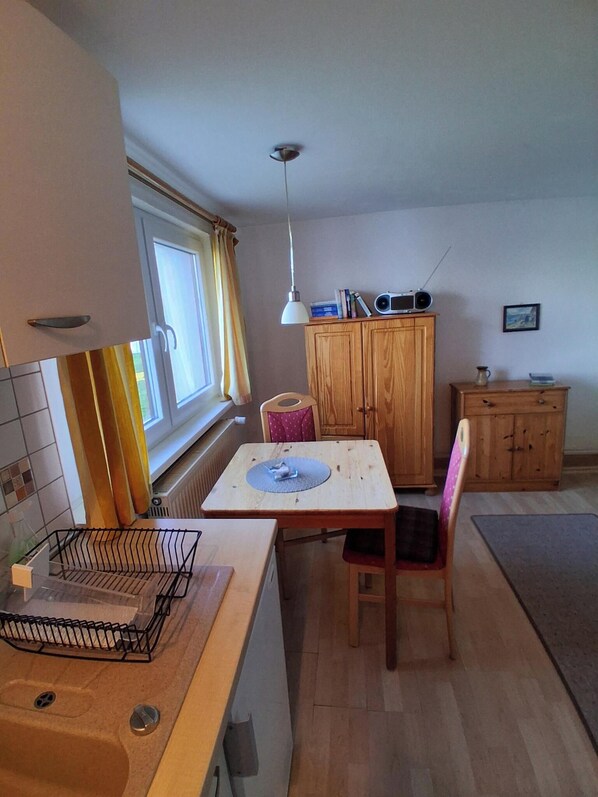 Une cuisine avec une table à manger, des meubles de rangement en bois, un luminaire suspendu et une fenêtre avec des rideaux.