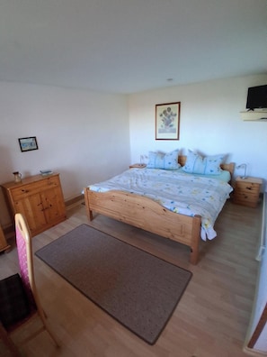 Une chambre avec un lit en bois, une commode en bois, une chaise, une petite table et un téléviseur fixé au mur.