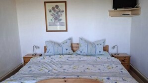 Ein hölzernes Bett mit floralem Bettbezug, zwei Nachttische, ein an der Wand montierter Fernseher und ein gerahmtes Kunstwerk.