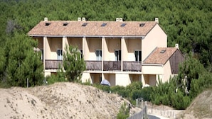 A multi-story building with balconies, surrounded by trees and a hilly landscape. - 3 BED Apartment With Garden (Carcans)