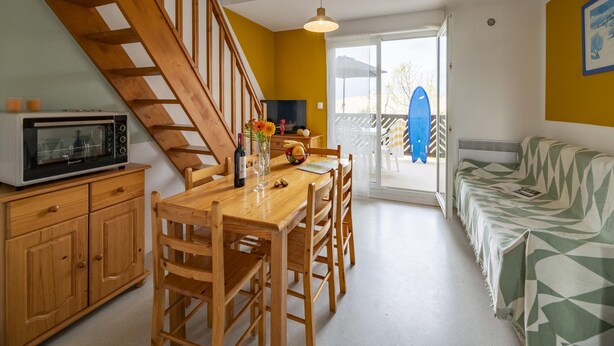 A kitchen with a wooden staircase, a dining table with chairs, a microwave, and a surfboard visible through the sliding glass door. - 3 BED Apartment With Garden (Carcans)