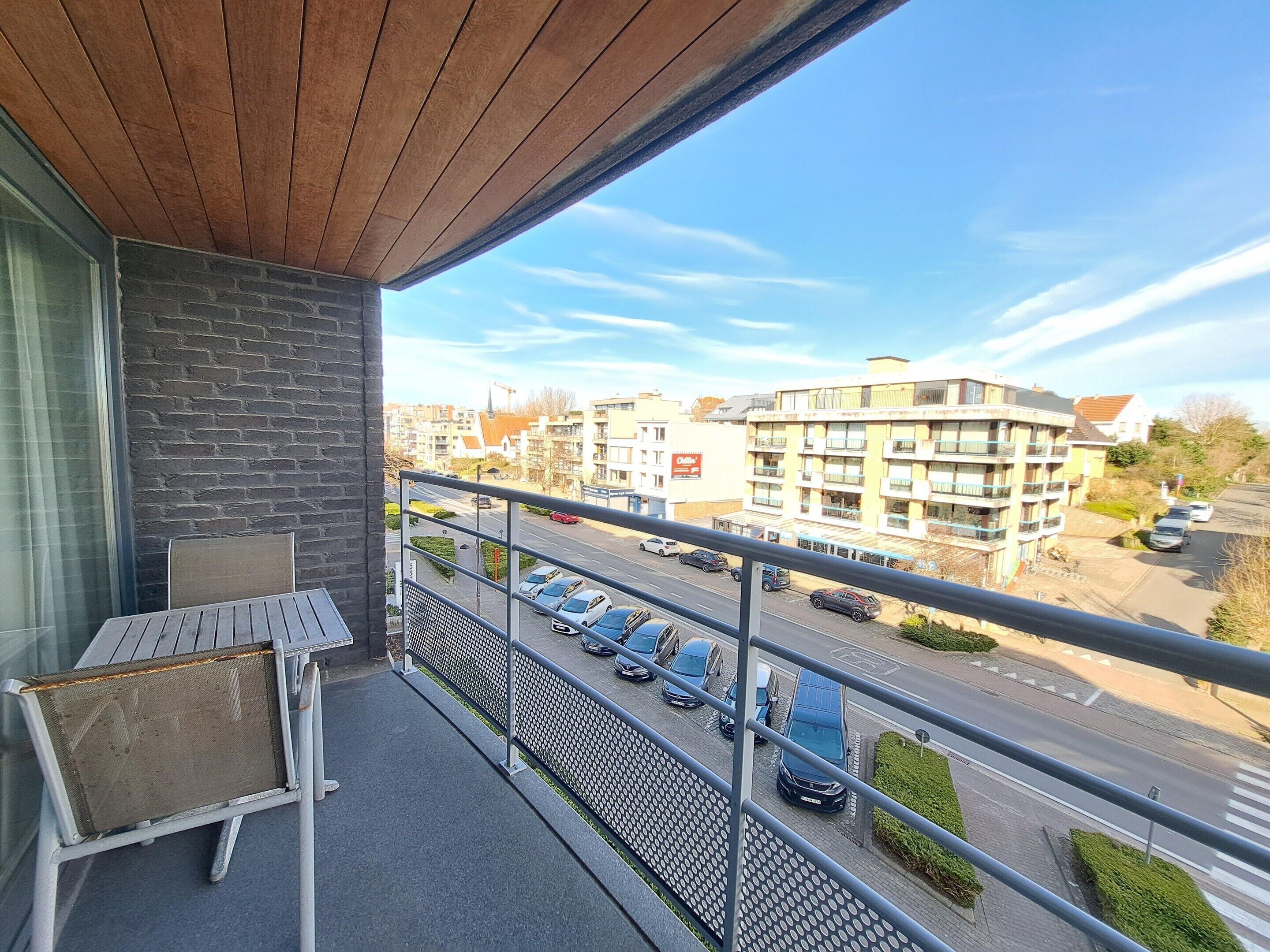 A balcony with a view of a street, a parked car, and a building with multiple balconies.