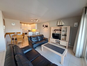 Ein modernes Wohnzimmer mit einer schwarzen Ledersofa, einem Couchtisch, einer TV-Konsole und einem Essbereich mit Stühlen, im Hintergrund befindet sich eine Küche.