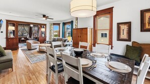 A dining area with a wooden table set for four, surrounded by white chairs, a chandelier, and a view of the outdoors through a window. - Walk to Downtown & Elkhart Lake | Firepit (Elkhart Lake)