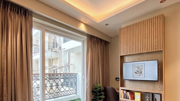 A modern bedroom with a bed, a desk, a chair, a TV on the wall, and a balcony with a railing. - Studio Ethereal by VoyZen Privé (Greater Noida)