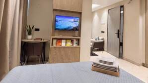 A hotel room with a bed, a TV mounted on a wooden panel, a desk with a chair, and a small potted plant. - Studio Ethereal by VoyZen Privé (Greater Noida)
