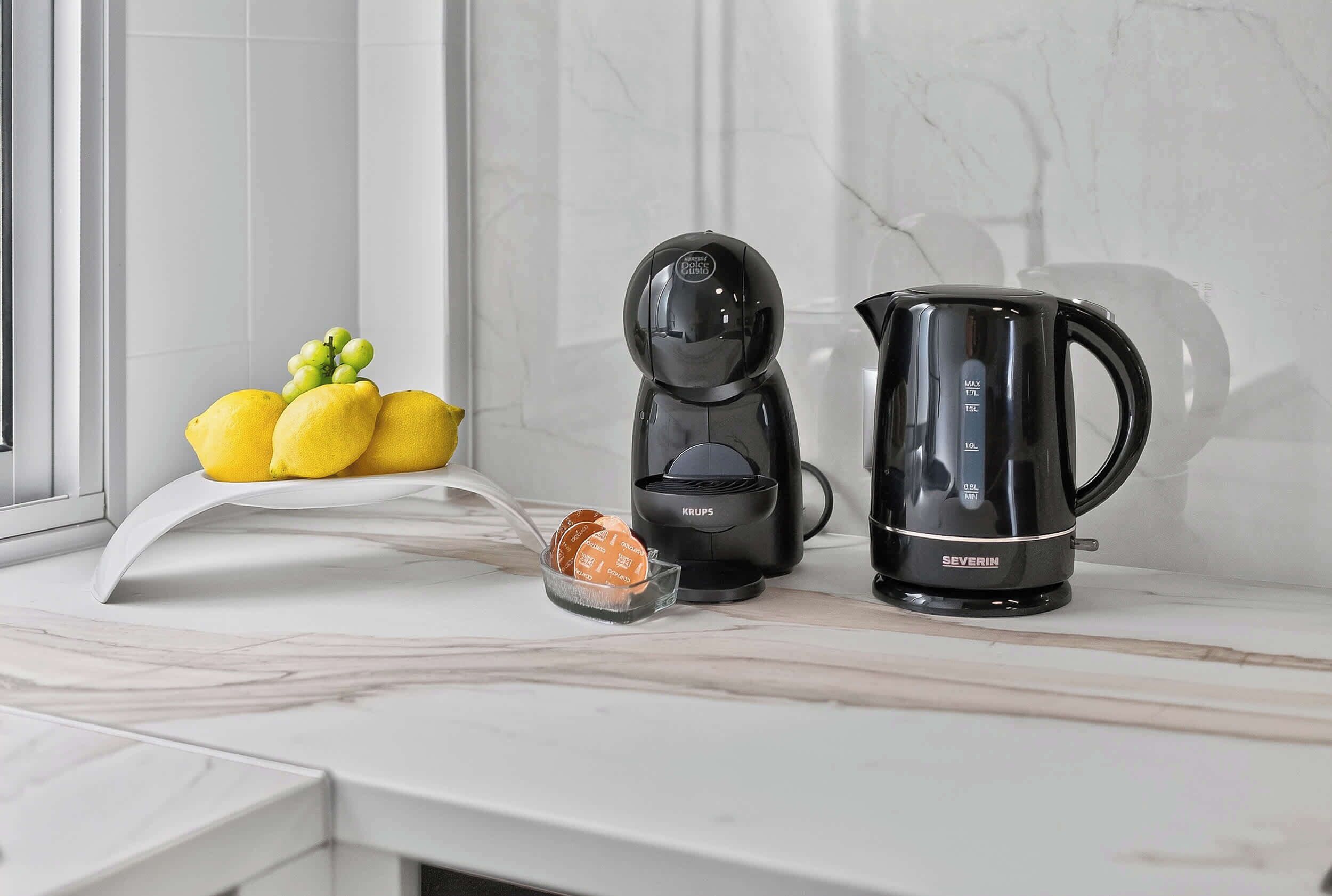 Una encimera de cocina con cafetera, hervidor y un cuenco de fruta.