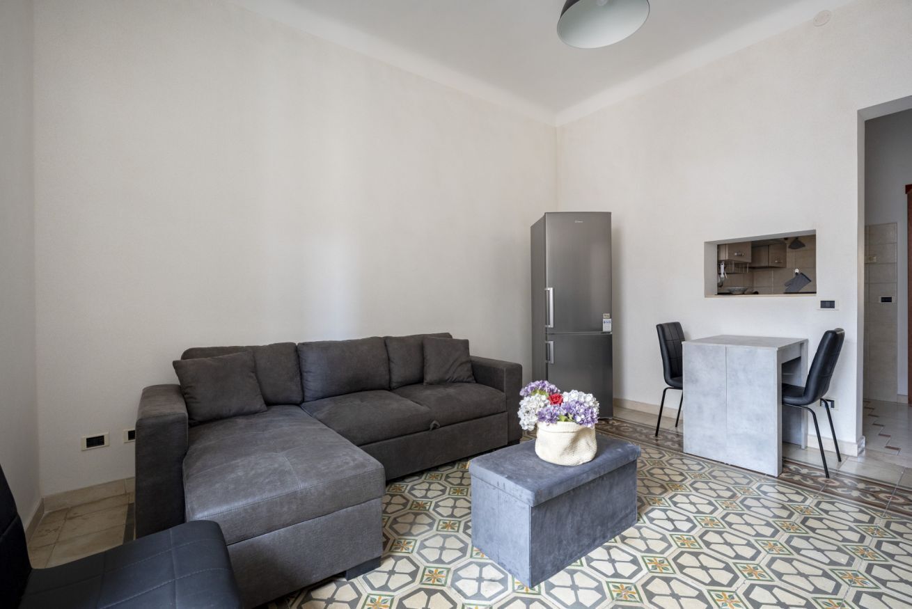 Uma sala de estar moderna com um sofá modular cinza, uma mesa pequena com duas cadeiras, um frigorífico cinza e uma área de cozinha visível através de uma porta aberta.