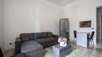 A modern living room with a grey sectional sofa, a small table with two chairs, a grey refrigerator, and a kitchen area visible through an open door.